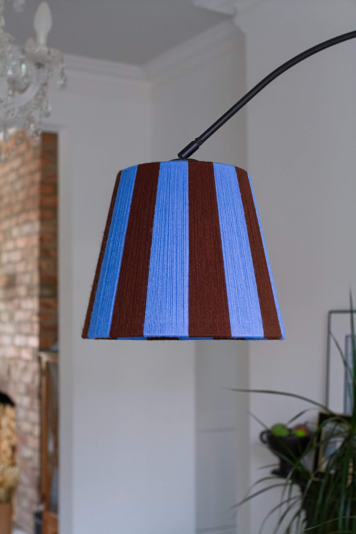 A close up of a modern floor lamp with a black arched stand and a slice shades yarn lampshade featuring vertical stripes of blue and brown styled in a room with a potted plant and cream curtains.