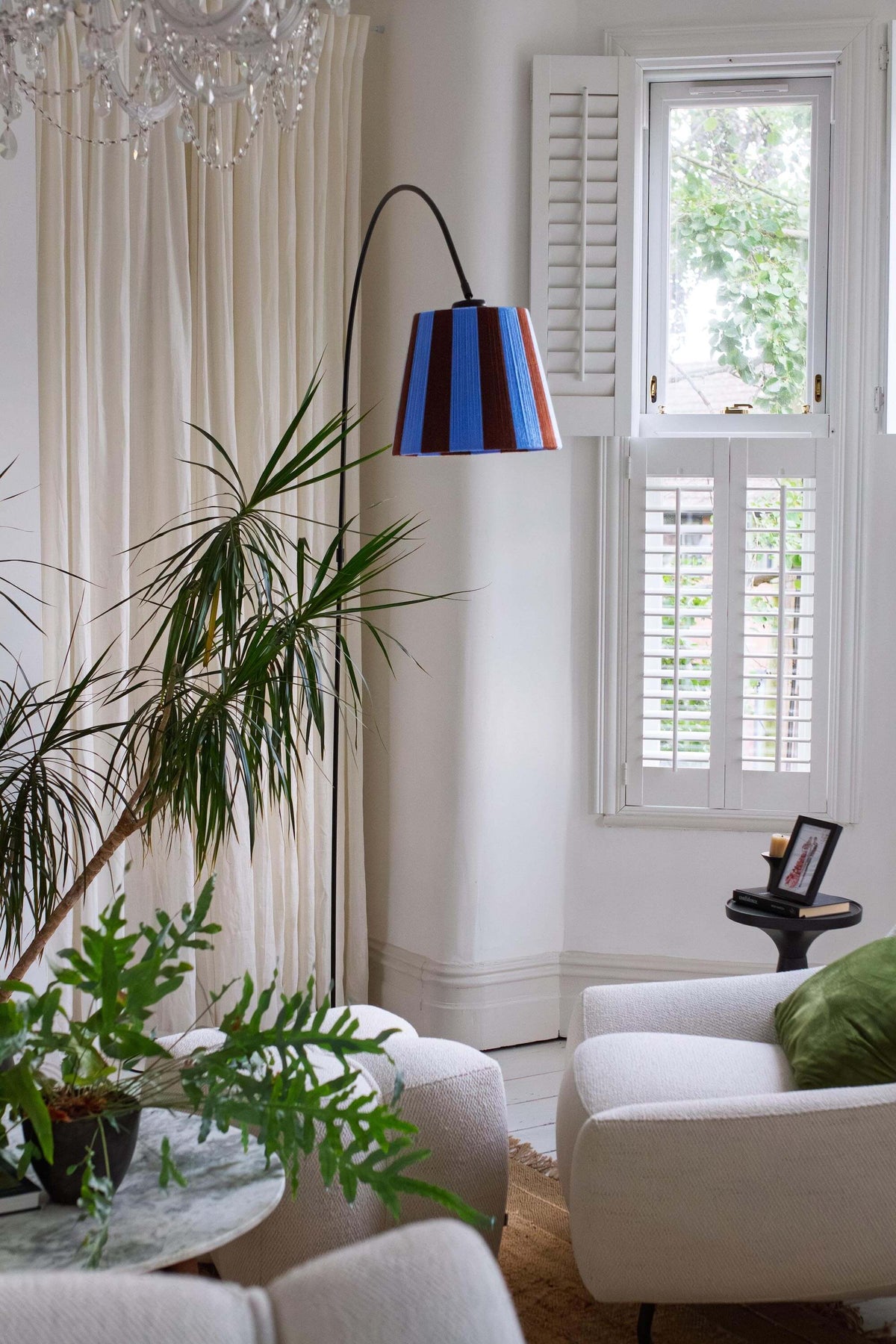 A modern floor lamp with a black arched stand and a slice shades yarn lampshade featuring vertical stripes of blue and brown styled in a room with a potted plant and cream curtains.