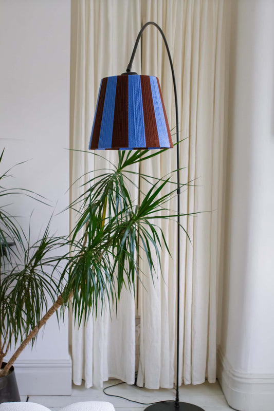 A modern floor lamp with a black arched stand and a slice shades yarn lampshade featuring vertical stripes of blue and brown styled in a room with a potted plant and cream curtains.