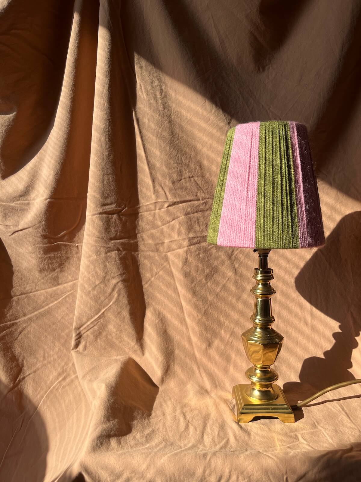 A small table lamp with a Slice Shades yarn lampshade withvertical stripes of pink and olive green, mounted on a polished brass base, set against a draped beige fabric backdrop with dramatic sunlight.