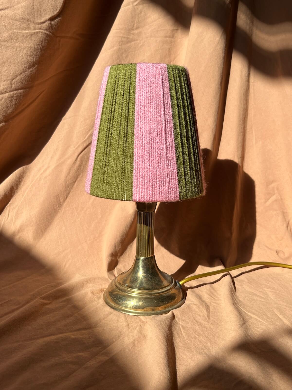 A vintage-style table lamp with a Slice Shades yarn lampshade featuring vertical stripes of olive green and pink, mounted on a polished brass stem and base, set against draped tan fabric with strong sunlight shadows.