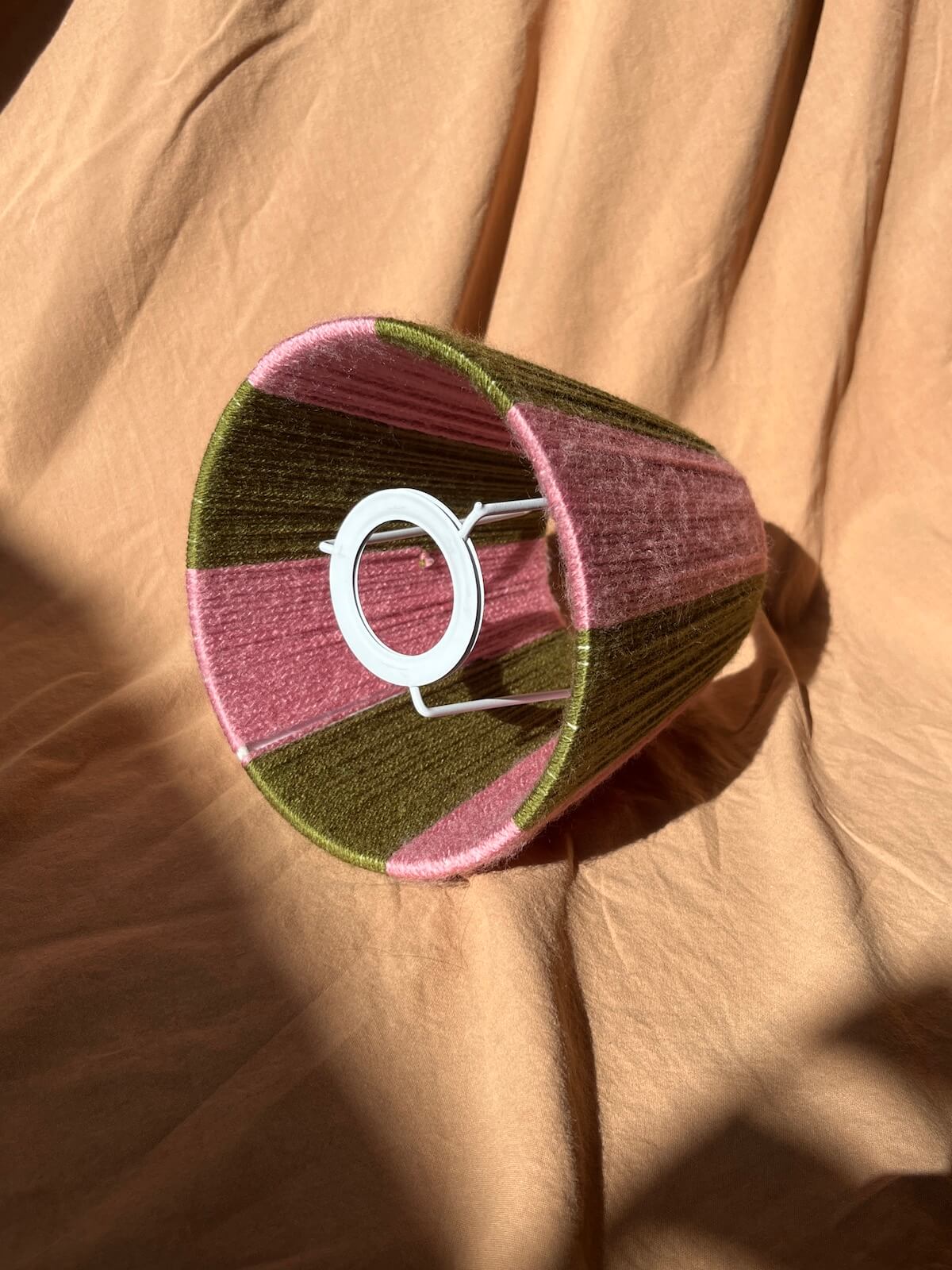 A handmade striped Slice Shades lampshade with alternating bands of dusty pink and olive green, displayed on a draped tan fabric background with strong natural sunlight and shadows.