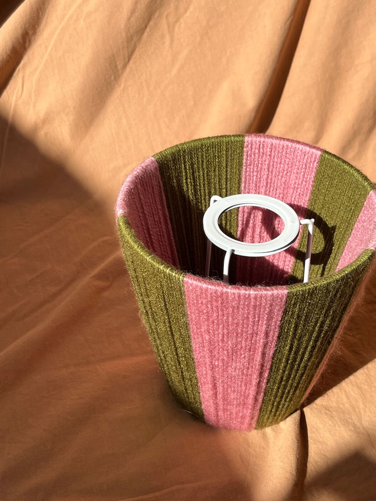 A Slice Shades lampshade with vertical stripes of olive green and dusty pink, set against a draped tan fabric background.