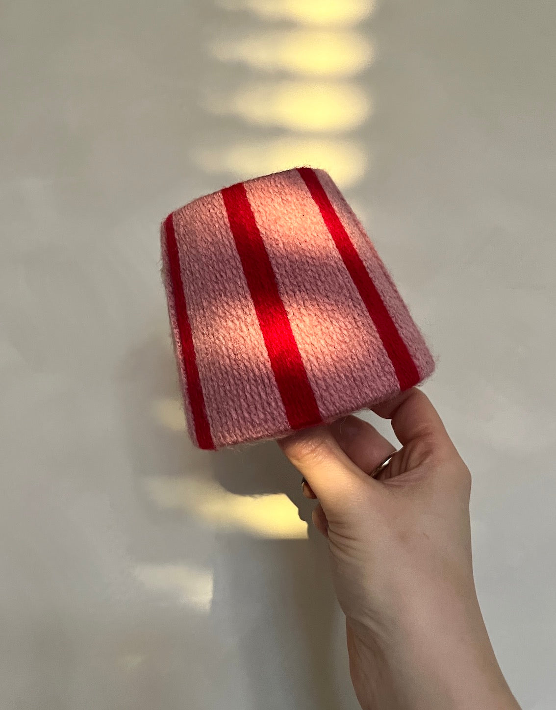 A hand holding a small, Slice Shades yarn lampshade with a pink and red striped pattern, set against a neutral light gray background with soft window light shadows.