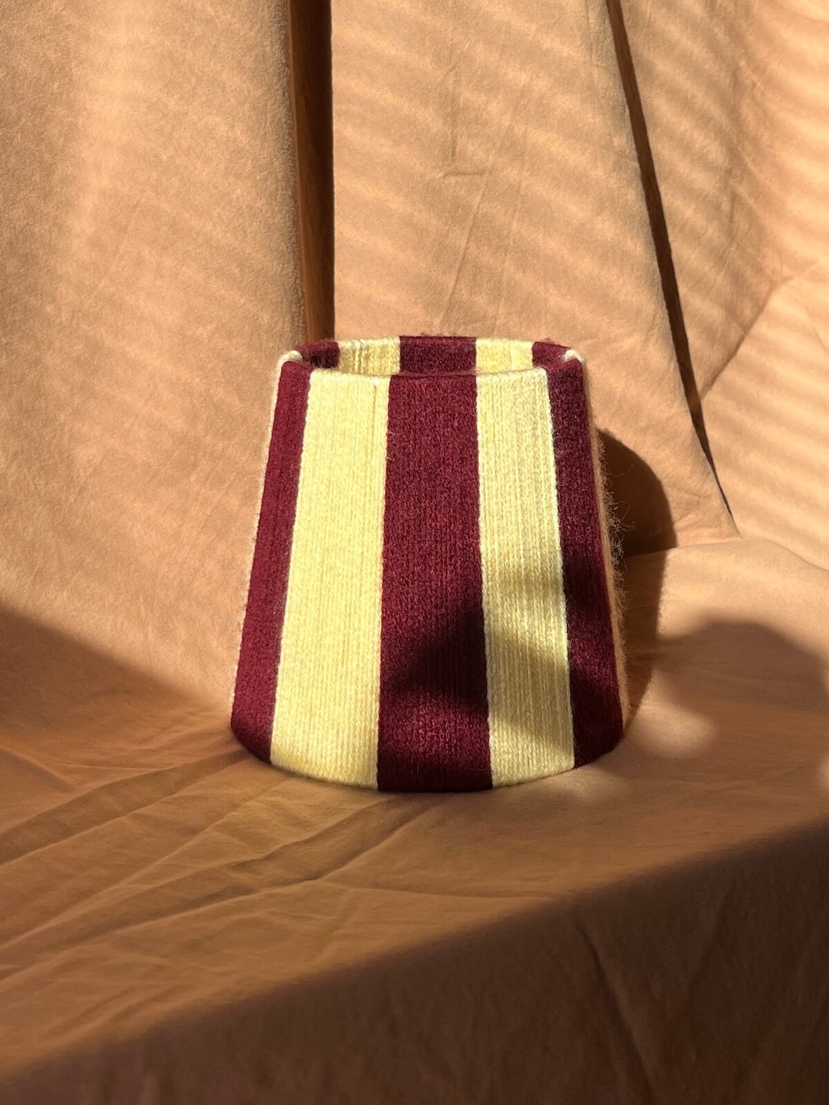 A mini string lampshade with vertical stripes in deep burgundy and butter yellow, set against a draped beige fabric backdrop with dramatic diagonal shadows.