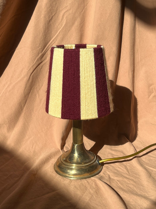 A small table lamp with a handowven string shade featuring vertical stripes in deep burgundy and butter yellow, mounted on a polished brass base with a visible power cord, set against a draped beige fabric background.