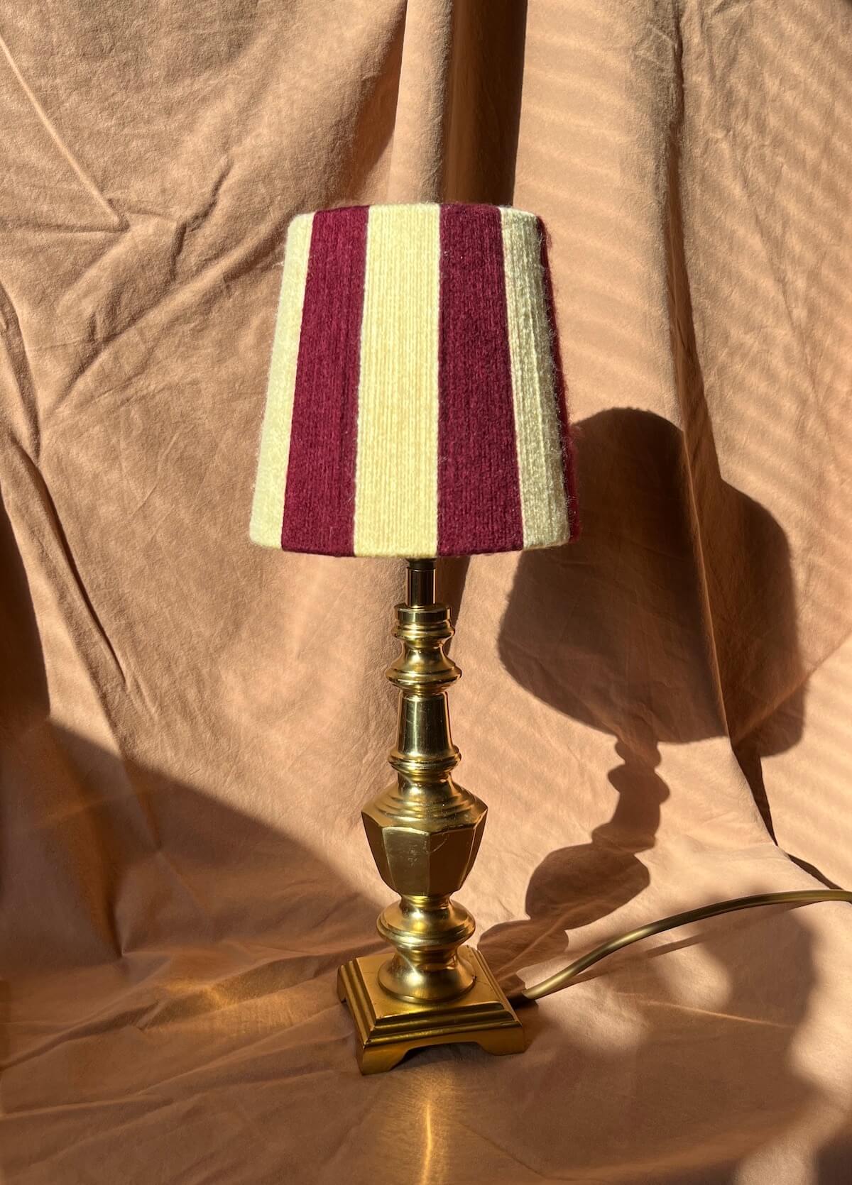 A table lamp with a striped yarn string shade in burgundy and butter yellow, mounted on a polished brass base, set against a draped beige fabric backdrop.