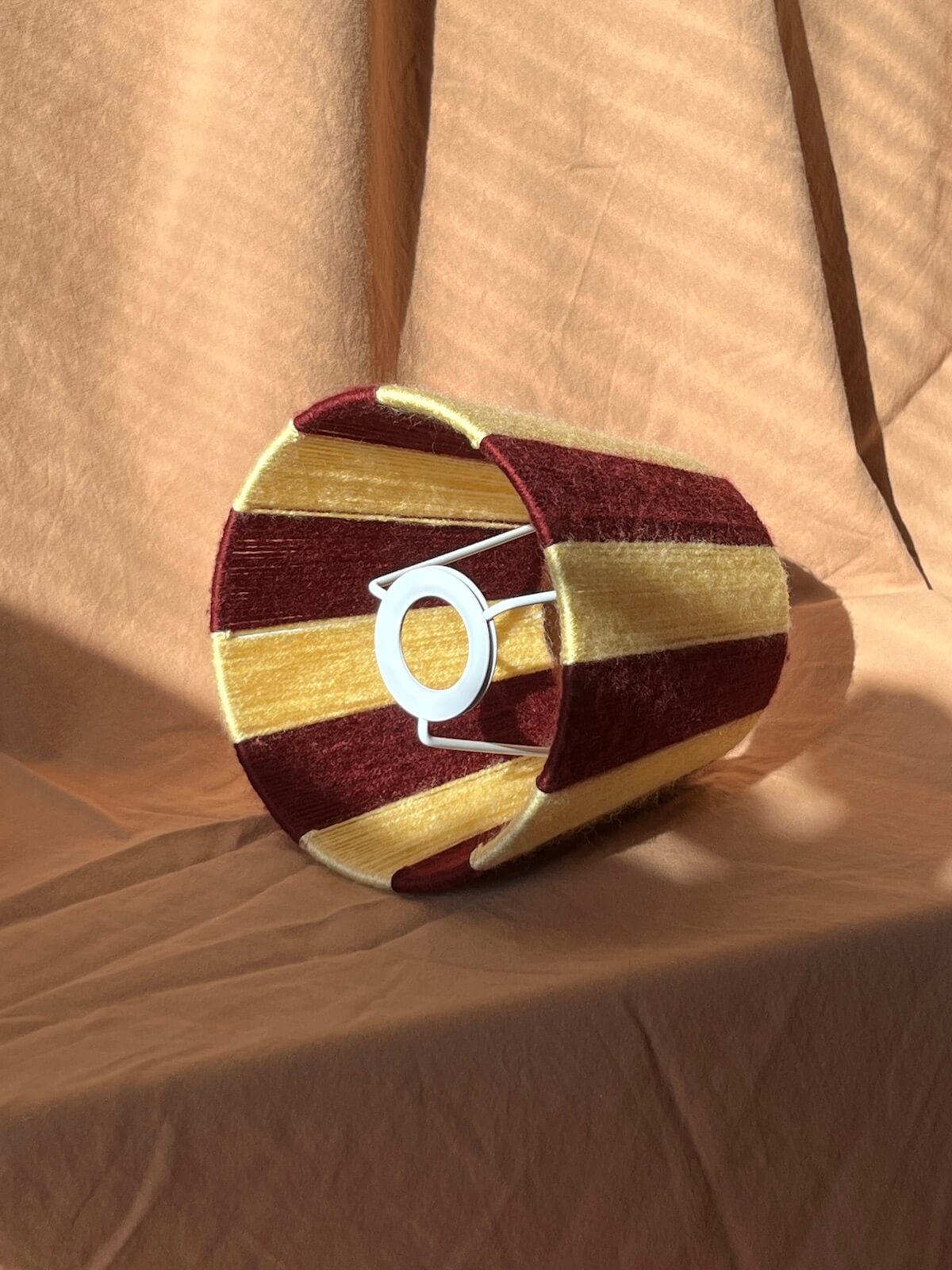 A string yarn lampshade with alternating horizontal stripes of deep burgundy and butter yellow with a fixed gimbal fitting, resting on draped tan fabric.