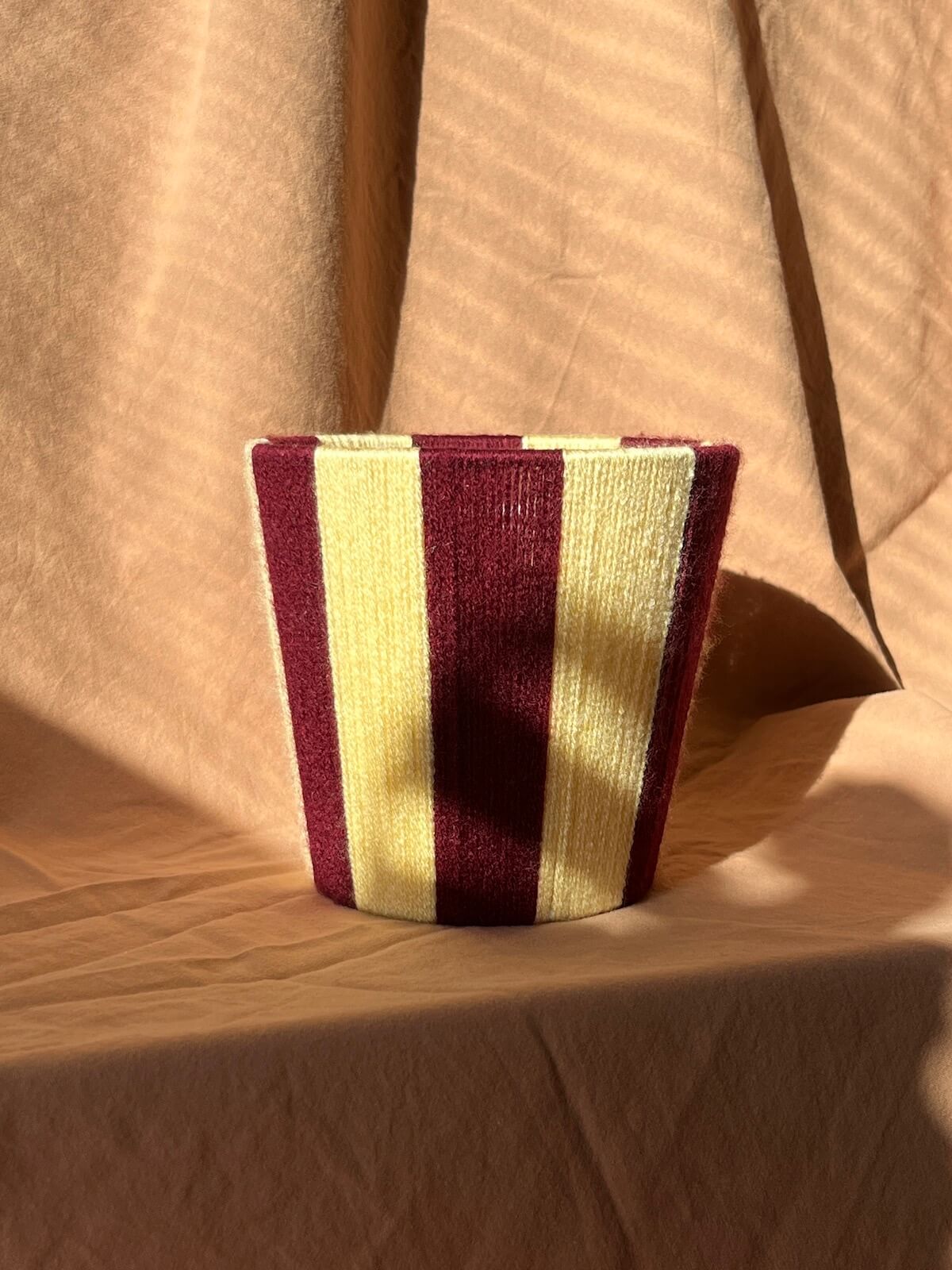 A small woven string lampshade with vertical stripes in deep burgundy and butter yellow, set against a draped beige fabric backdrop with strong sunlight and shadows.