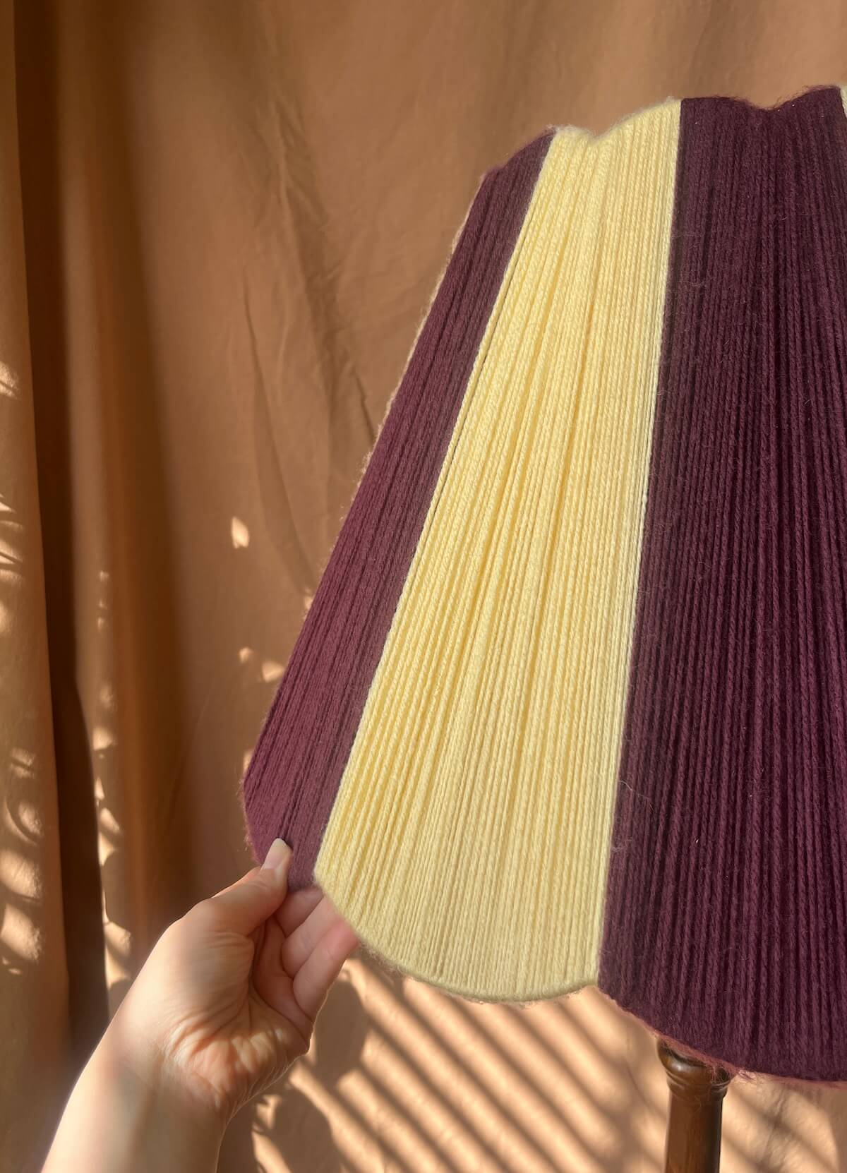 A Slice Shades scallop yarn lampshade held by a hand, featuring three vertical panels in butter yellow and plum set against a brown draped fabric background with leaf shadows.