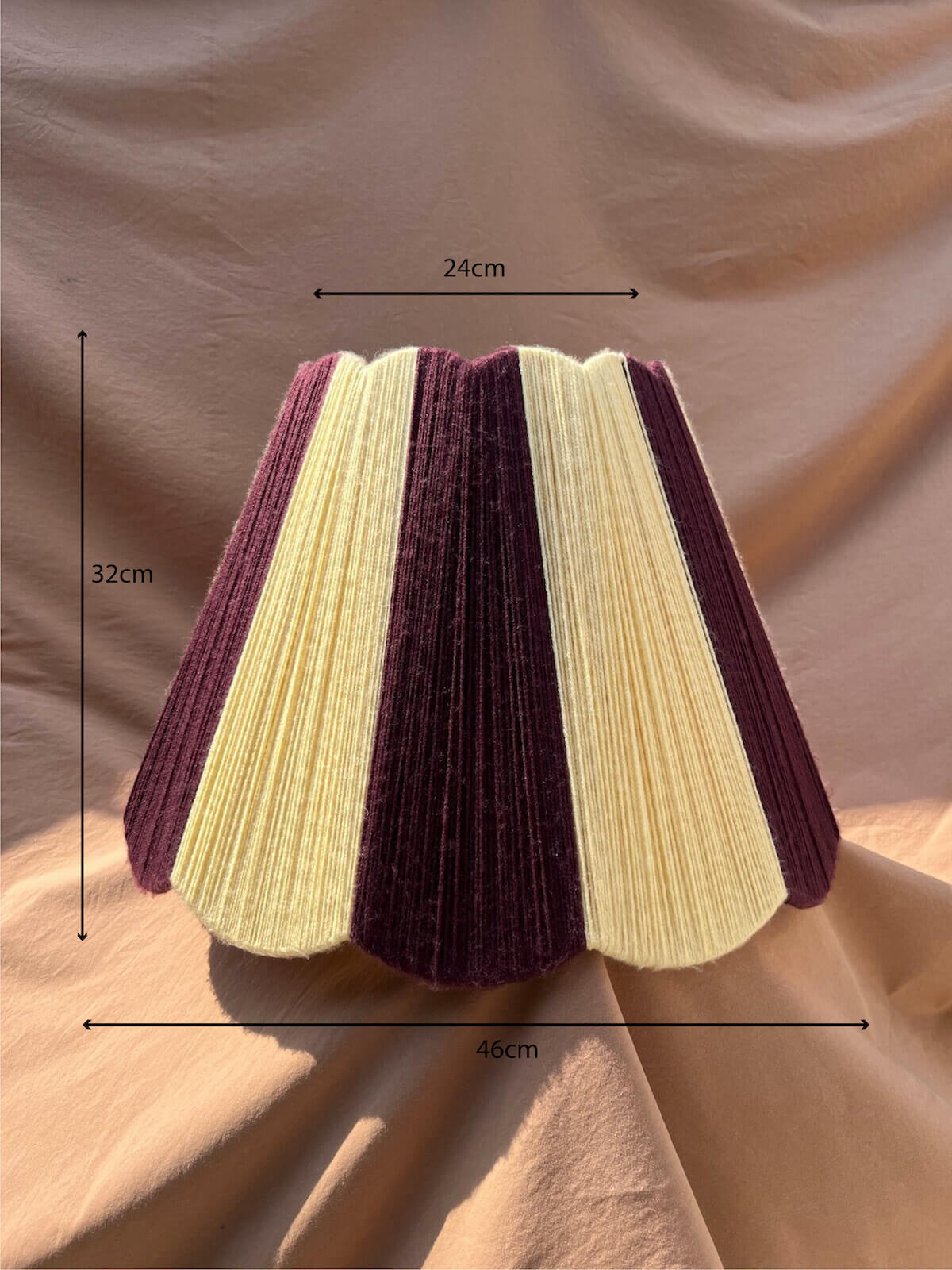 A Slice Shades Scallop Yarn lampshade with alternating vertical stripes of deep plum purple and yellow, displayed against a draped beige fabric backdrop. Shown with black dimension lines and text indicating its height of 32cm, width of 24cm, and base diameter of 46cm, set against a draped beige fabric background.
