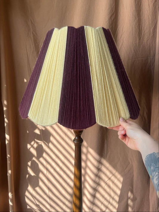 A Slice Shades Scallop Yarn lampshade in alternating vertical stripes of deep purple and pale yellow, mounted on a dark wooden stand. A hand is holding the right side of the shade.