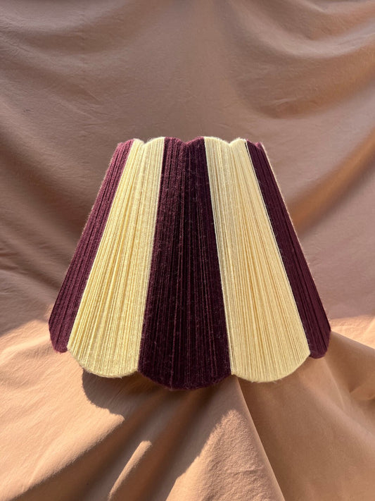 A Slice Shades Scallop Yarn lampshade with alternating vertical stripes of deep plum purple and yellow, displayed against a draped beige fabric backdrop.