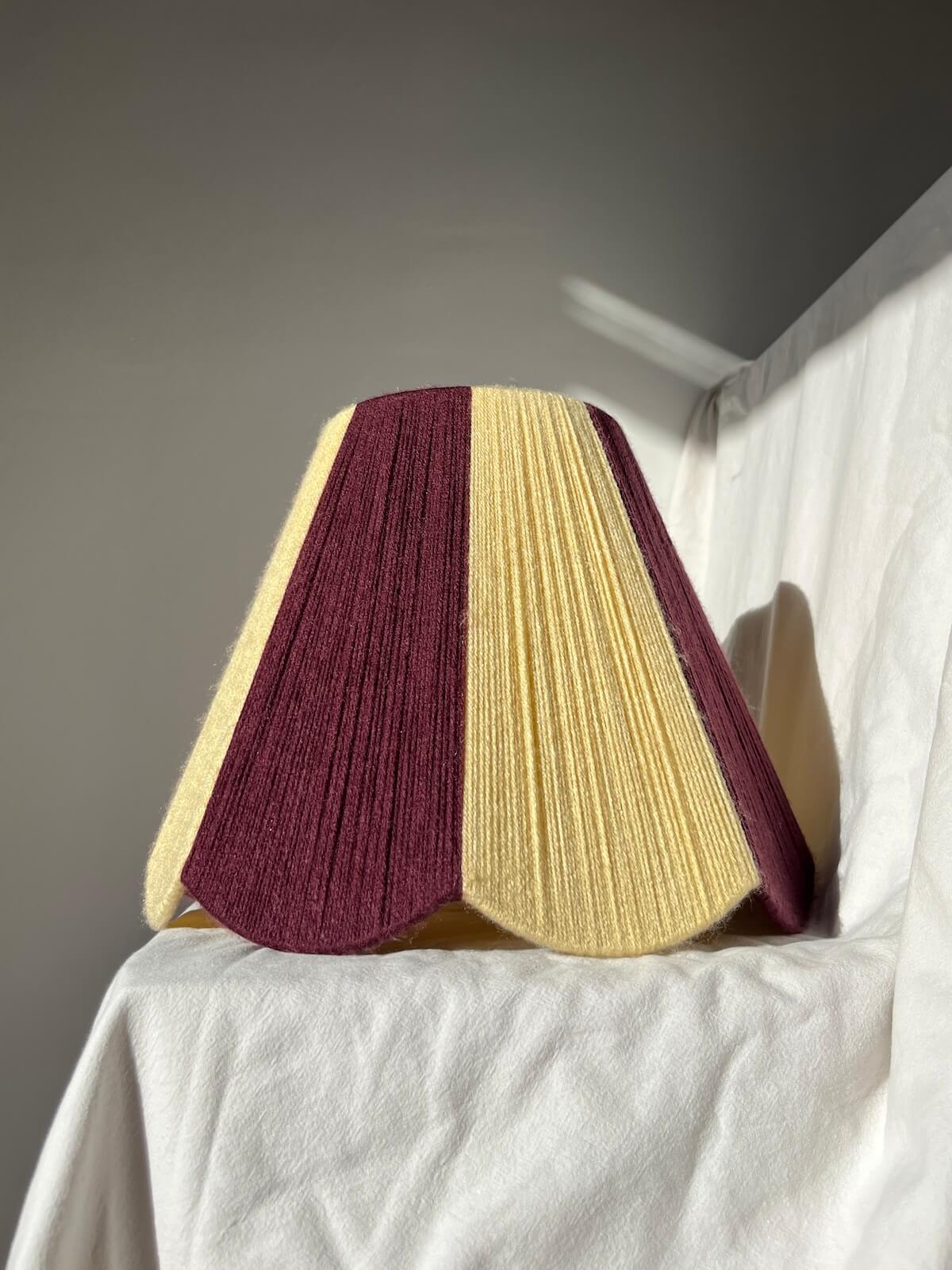 A Slice Shades lampshade with a handwoven yarn design of textured buttermilk yellow and deep burgundy, displayed on a draped white fabric background.