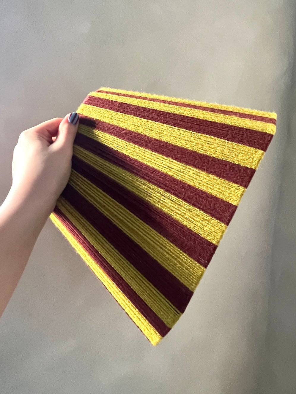 A hand holding a Slice Shades handwoven yarn lampshade with alternating horizontal stripes of dark burgundy and golden chartreuse, set against a plain light gray background.