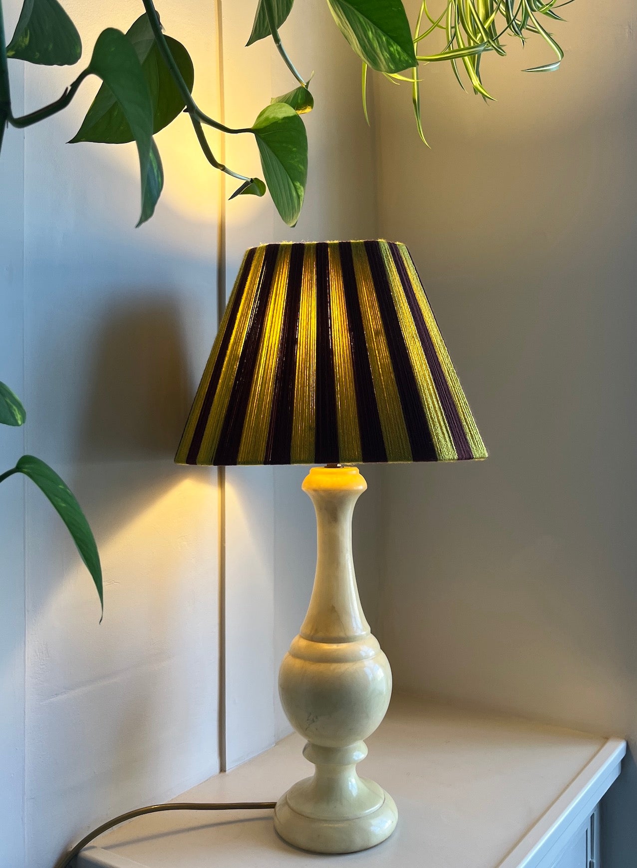 A table lamp with a cream-colored turned wood base and a Slice Shades lampshade featuring alternating mulberry and mustard yellow stripes, illuminated on a tabletop with houseplant leaves in the foreground.