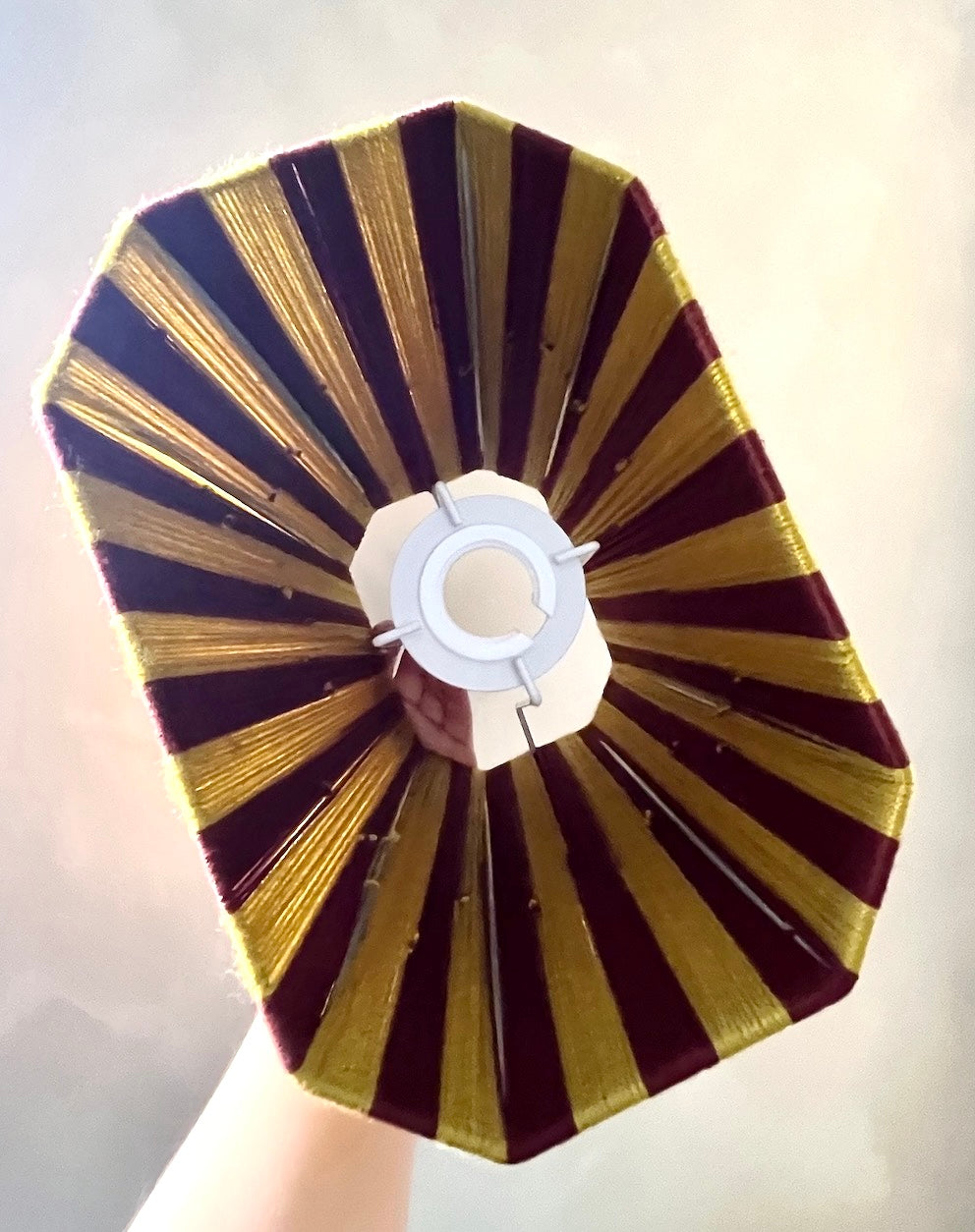 A hand holding a Slice Shades octagonal lampshade featuring alternating radiating stripes of deep burgundy and chartreuse gold thread, with a white fabric centre band.
