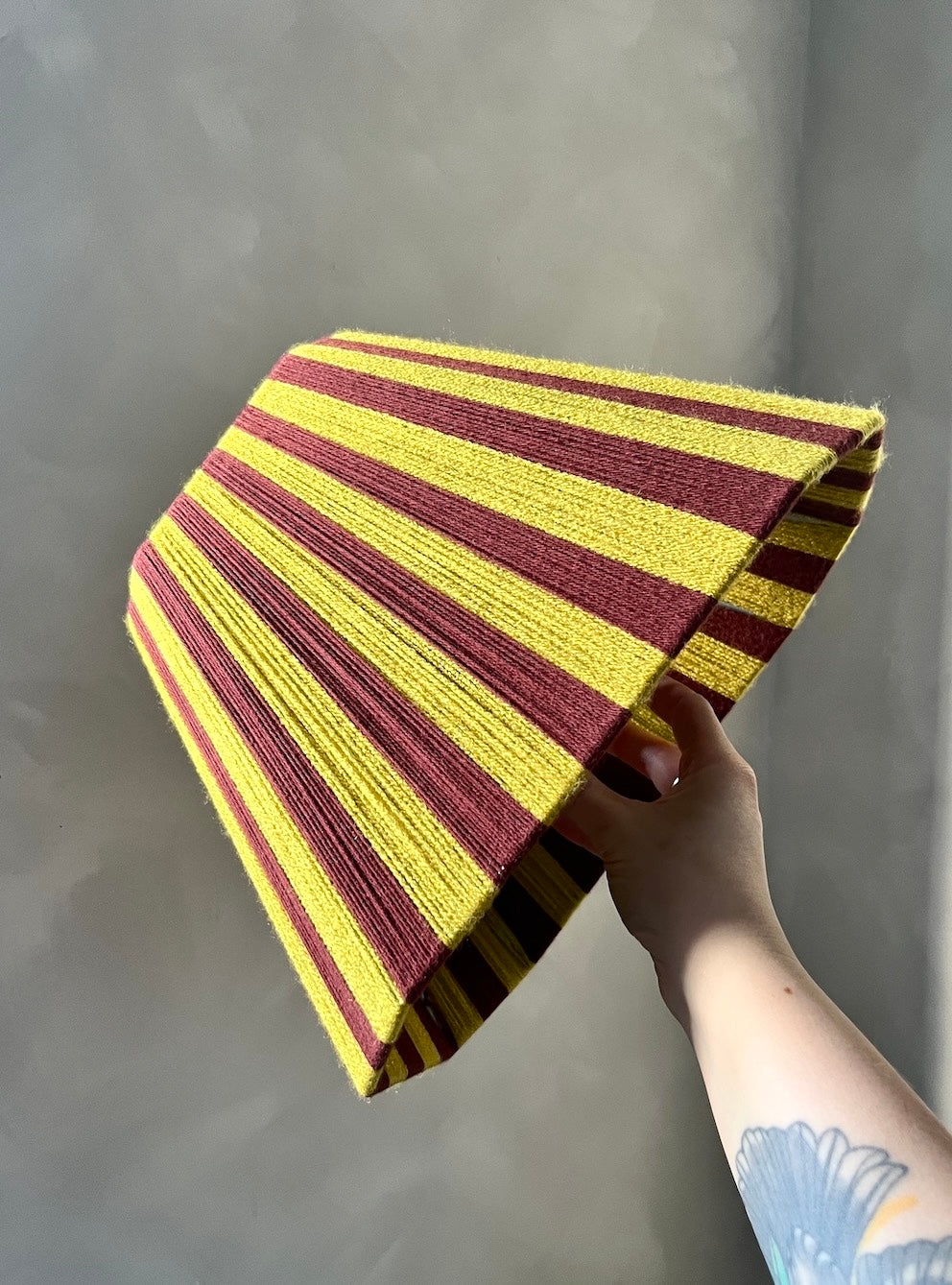 A hand holding a striped handmade Slice Shades yarn lampshade with alternating yellow and dark red stripes, set against a plain gray wall.
