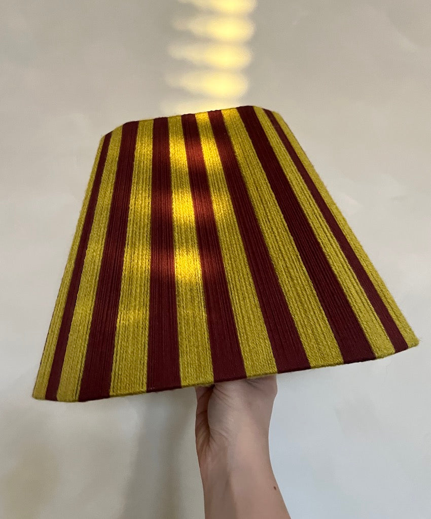 A hand holding a handmade yarn Slice Shades lampshade with vertical stripes in chartrese and dark red, set against a plain light background.