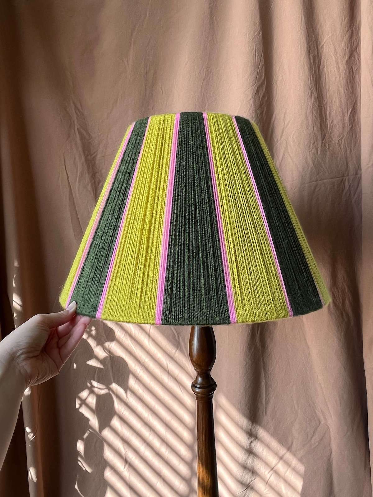 A Slice Shades yarn lampshade with vertical stripes of lime green, dark green, and pink, shown on a wooden floor lamp with a hand holding the left edge against a brown curtain backdrop.