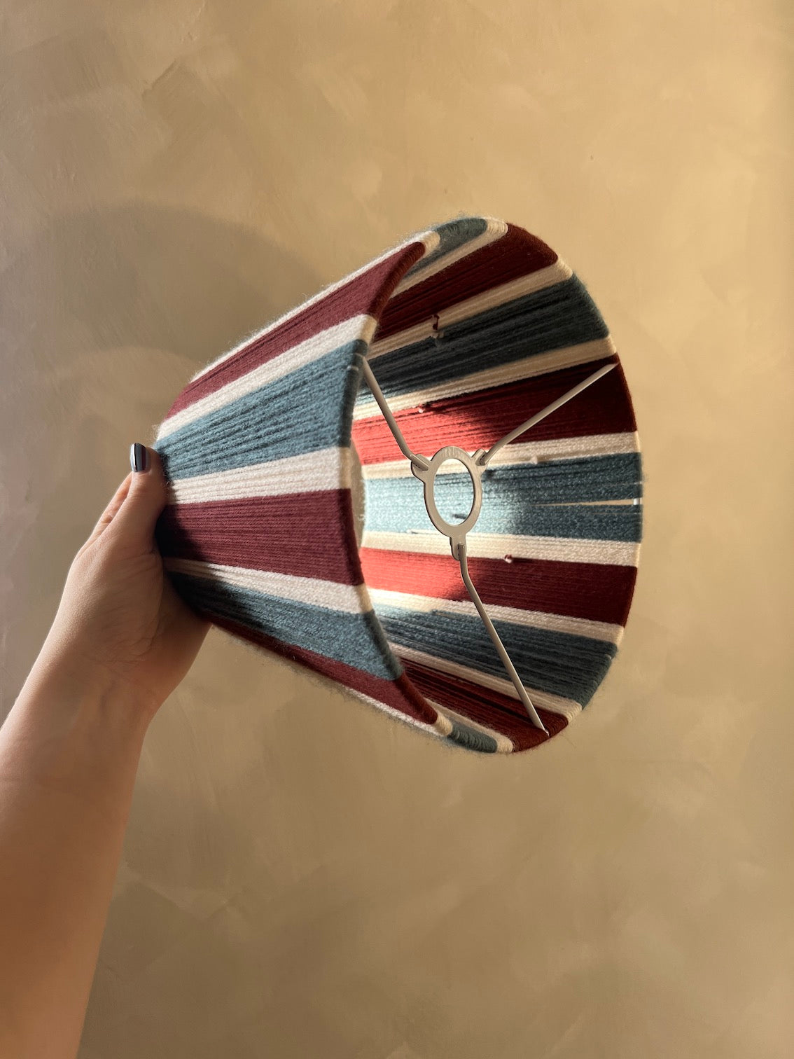 A hand holding a striped Slice Shades lampshade with vertical stripes in burgundy  white, and blue, against a plain beige wall.