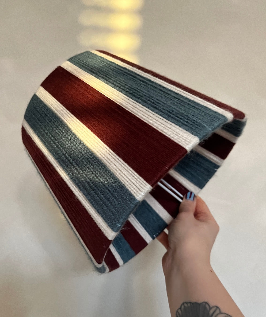 A striped Slice Shades lampshade held by a hand. The shade features alternating stripes of dark red, slate blue, and white.