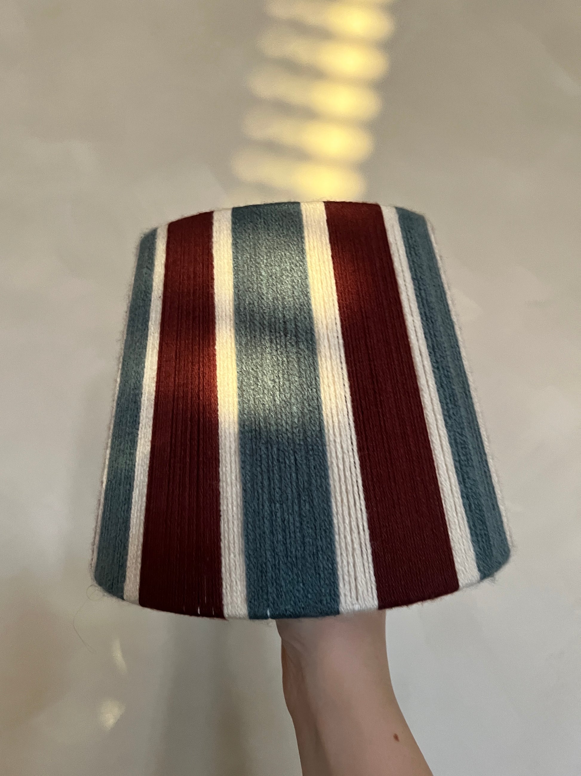 A hand holding a Slice Shades lampshade with vertical stripes in burgundy, slate blue, and cream, set against a warm off-white wall with soft window light shadows.