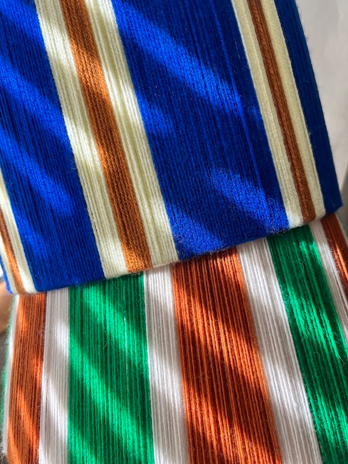 Close-up of striped patterns on Slice Shades yarn lampshades with vertical bands of blue, orange, cream, and green under dappled sunlight.
