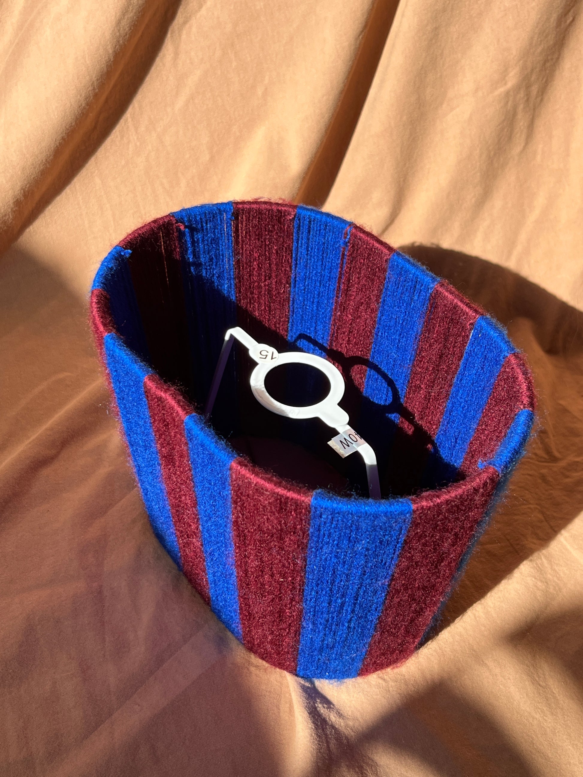A striped yarn oval lampshade in alternating blue and burgundy stripes, shown with fixed gimbal, placed on beige fabric under direct sunlight.