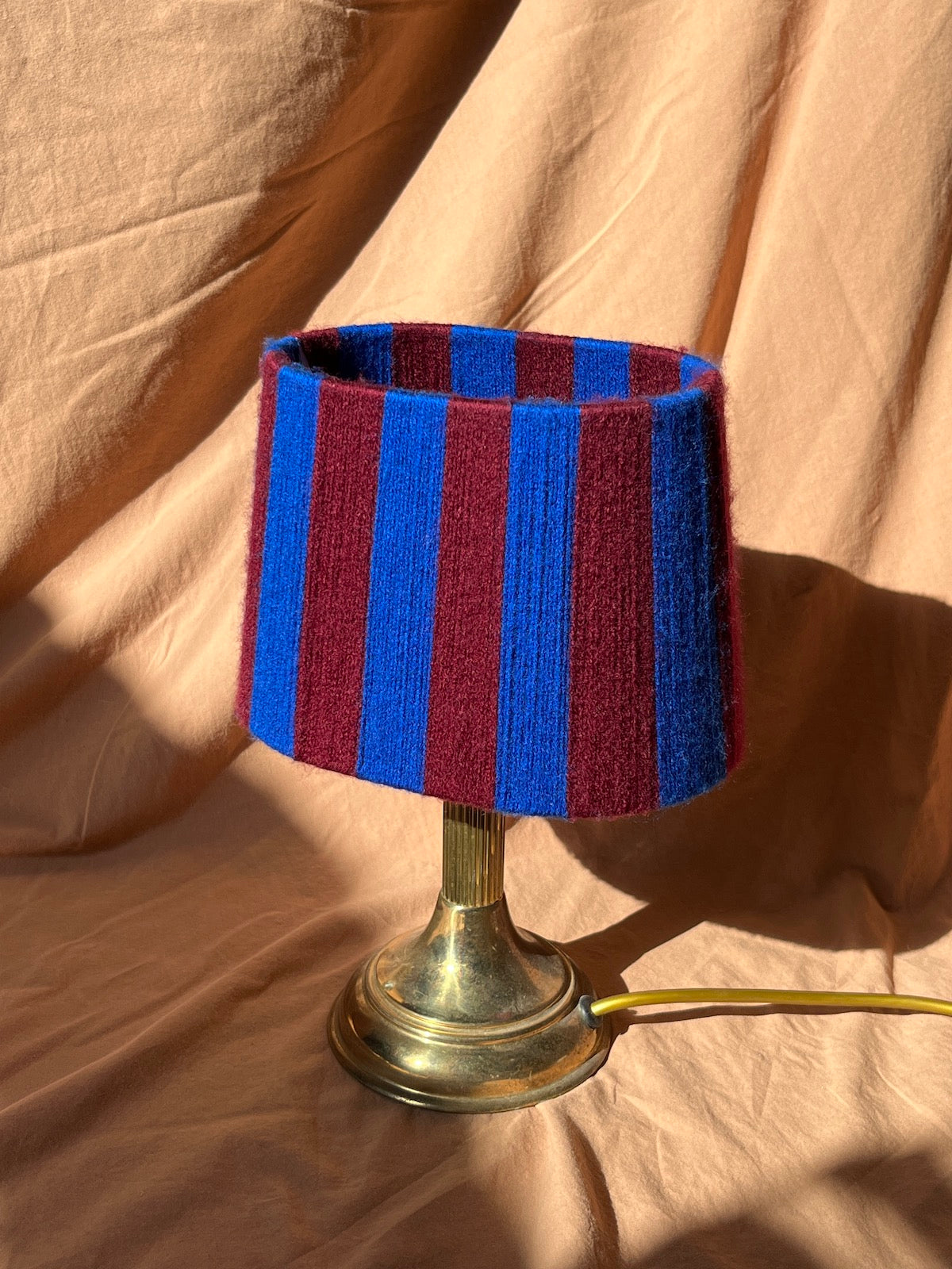 A table lamp with a n oval string yarn lampshade featuring vertical stripes of cobalt blue and deep burgundy, mounted on a vintage brass-colored metal base with a visible cord.