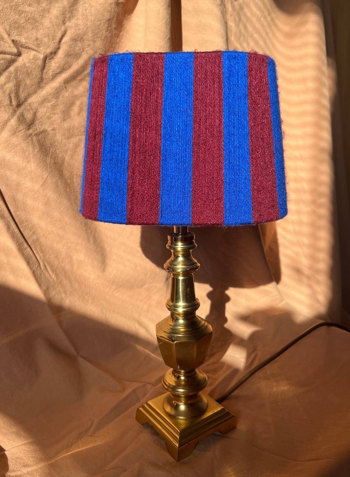A table lamp with a striped yarn oval lampshade in alternating blue and burgundy stripes, mounted on a polished brass-coloured metal base with a square footed stand, set against a draped beige fabric backdrop.