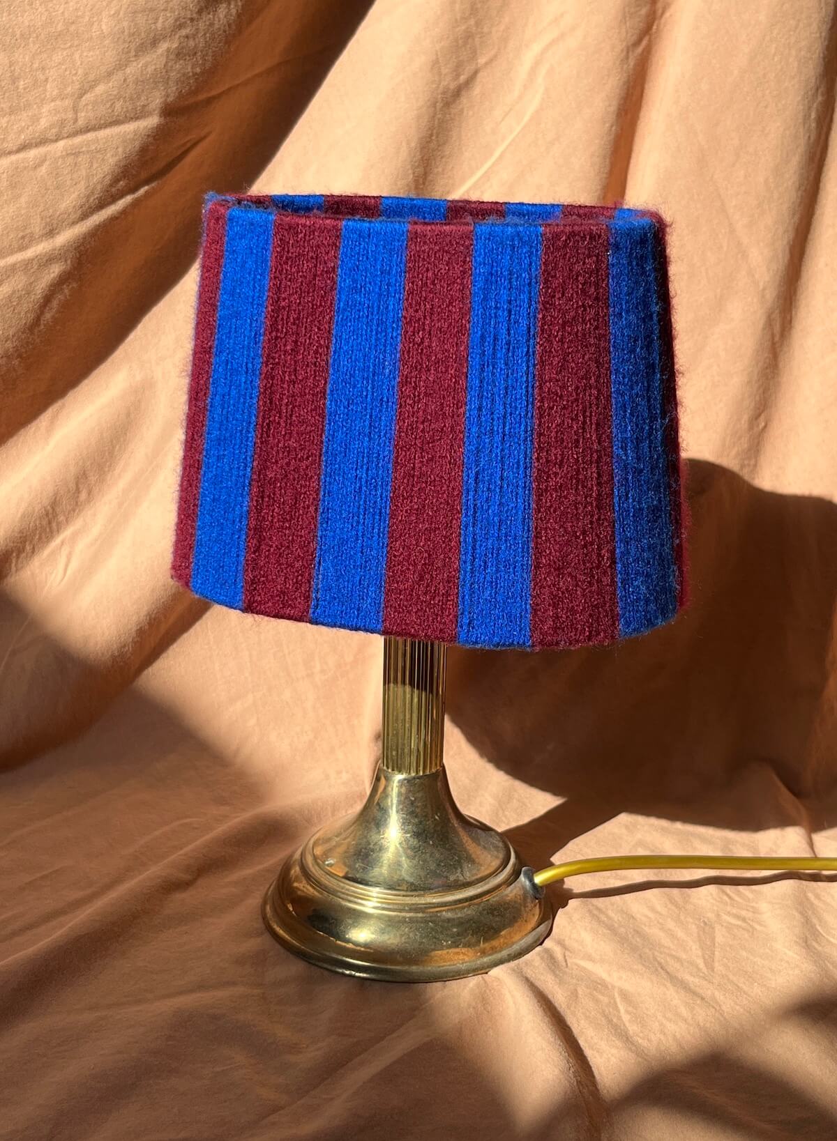 A table lamp with a striped yarn oval lampshade in alternating blue and burgundy stripes mounted on a metallic gold-toned base, set against a draped beige fabric backdrop.