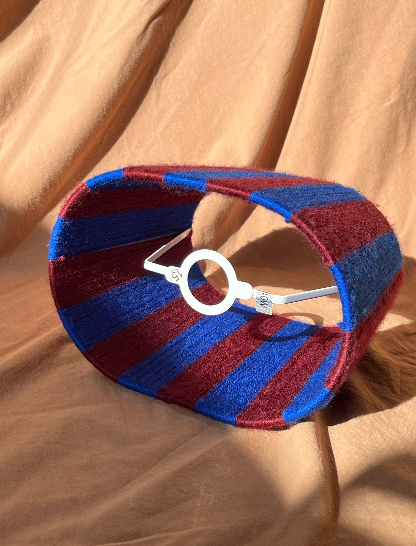 A striped yarn oval lampshade in alternating blue and burgundy stripes side view showing fixed set against a beige fabric backdrop.