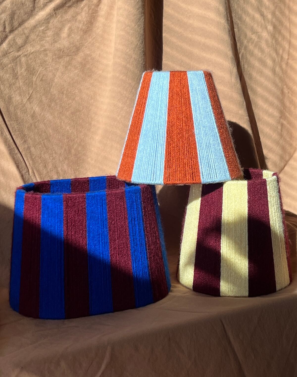 A striped yarn oval lampshade in alternating blue and burgundy stripes amongst other woven stripey lampshades set against a beige fabric backdrop.