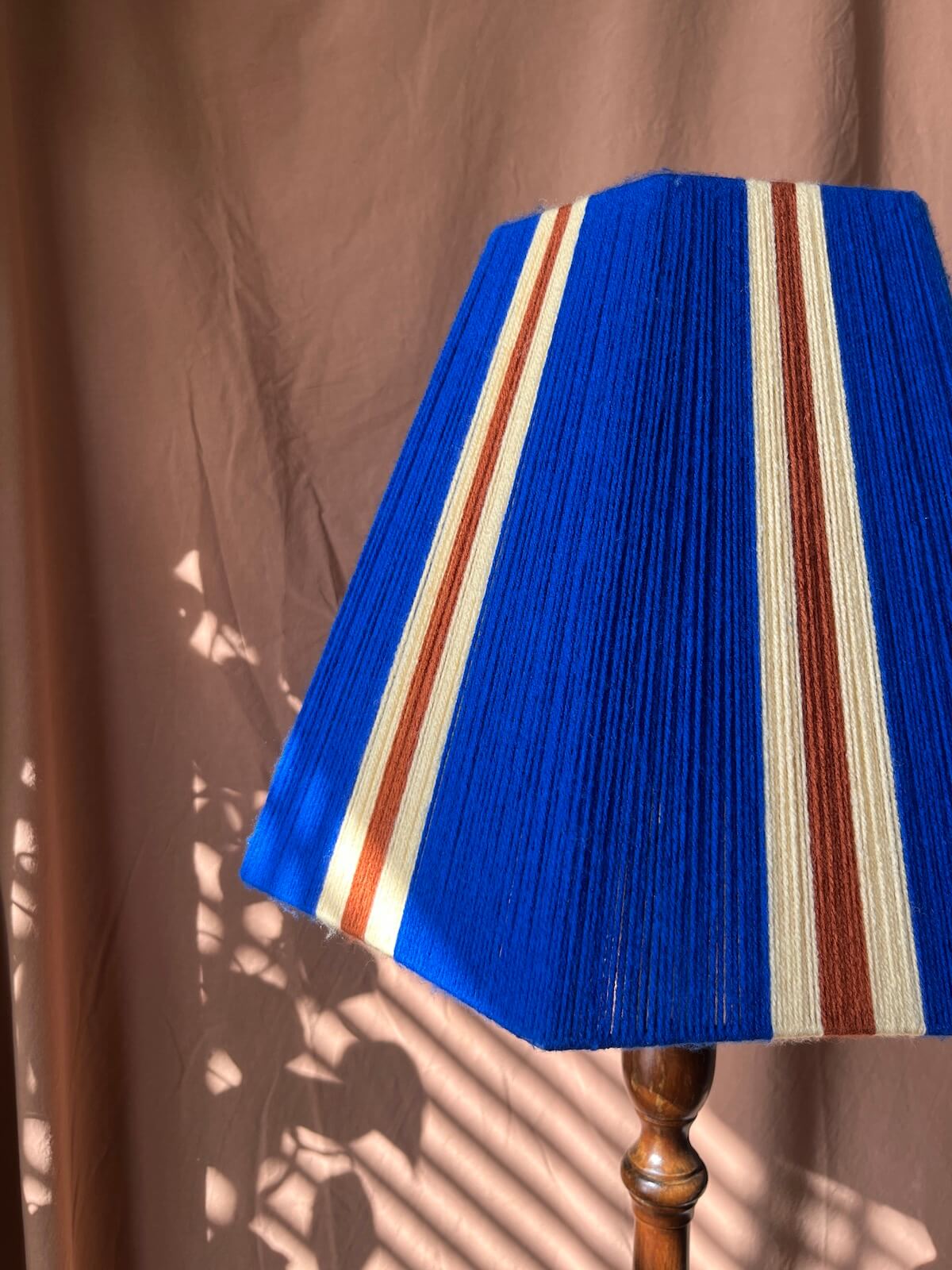 A Slice Shades yarn lampshade with vertical stripes in royal blue, lemon and rust red, mounted on a wooden lamp stand against a draped brown fabric backdrop with leaf shadows.