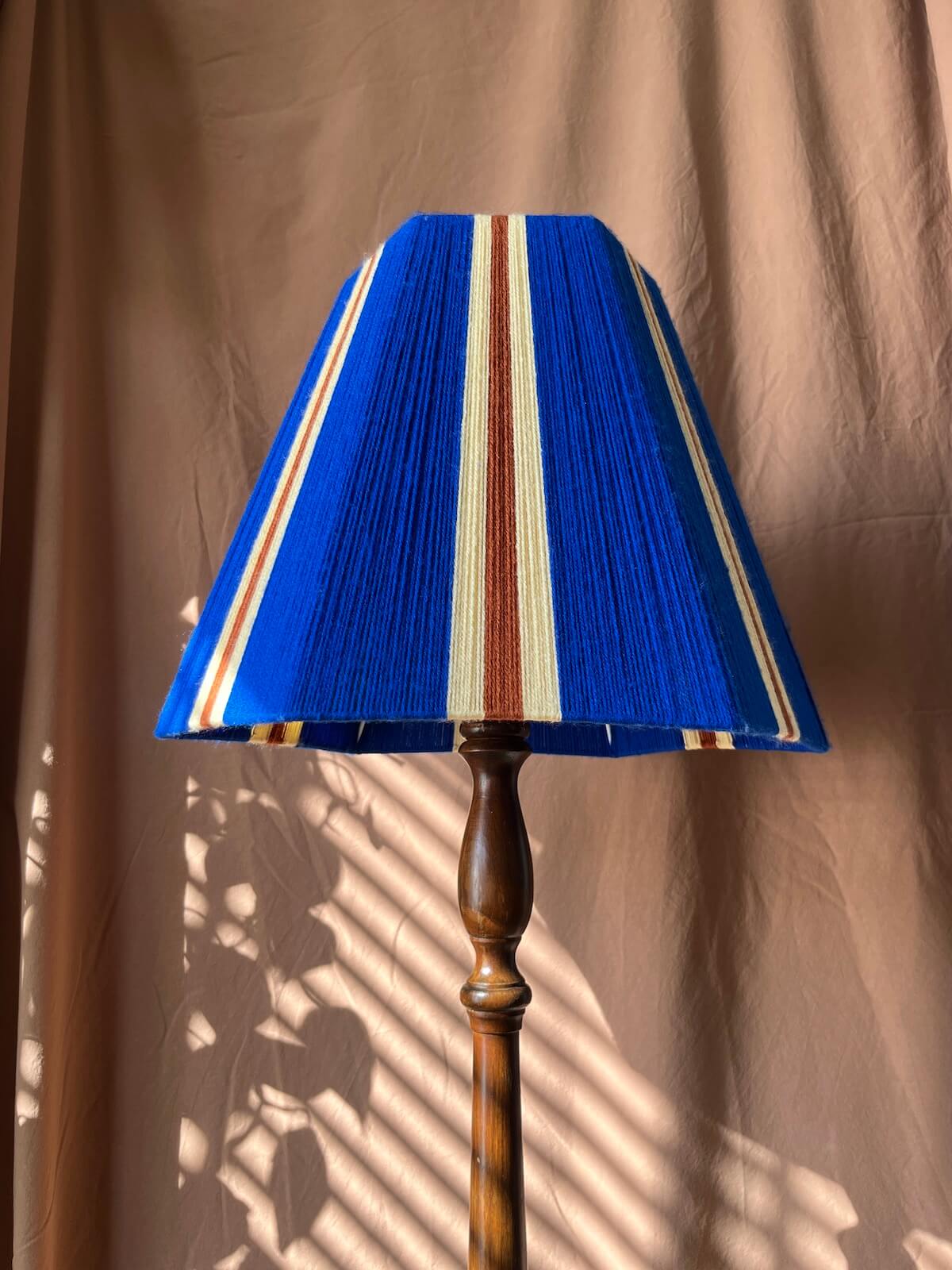 A Slice Shades yarn lampshade in royal blue with vertical yellow and rust-orange stripes, mounted on a dark brown wooden floor lamp stand against a draped tan fabric backdrop with dappled sunlight shadows.
