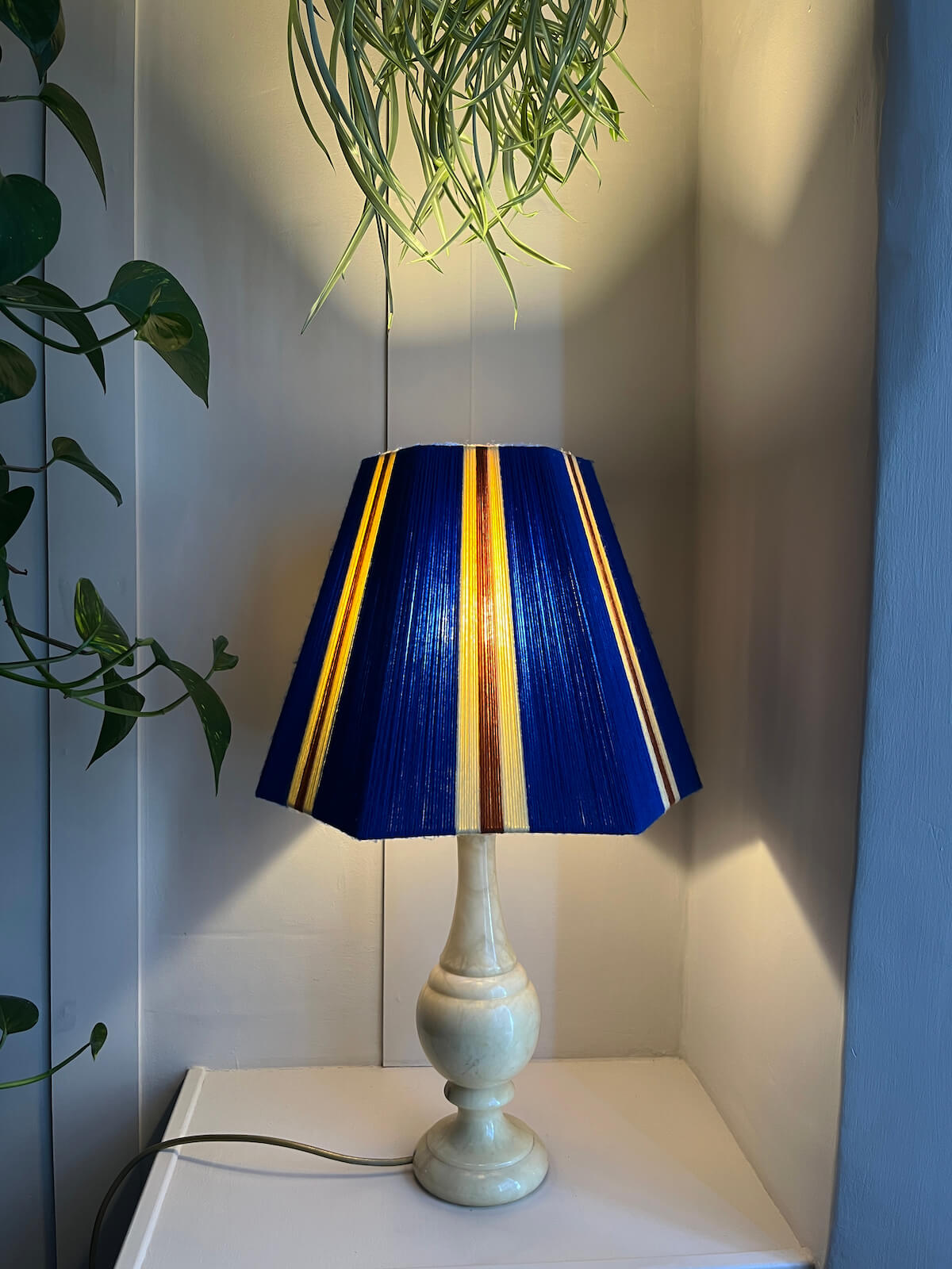 A Slice Shades yarn lampshade in royal blue with vertical yellow and rust-orange stripes, set on a white side table against a panelled wall with houseplants glowing.