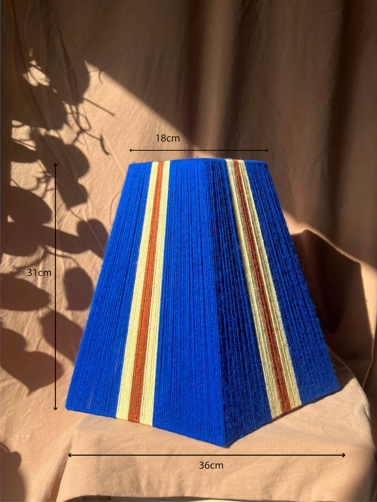 A Slice Shades yarn lampshade in a vibrant royal blue, featuring two vertical stripes of lemon yellow and rust brown, displayed on a beige fabric backdrop with leaf shadows cast across it with dimensions