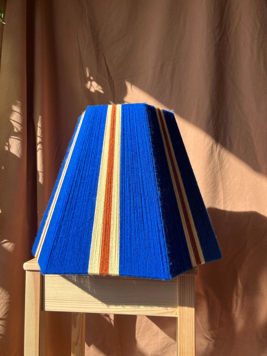 A Slice Shades yarn lampshade with vertical stripes in lemon, orange, and cobalt blue, displayed on a light wood stool against a draped tan fabric backdrop.