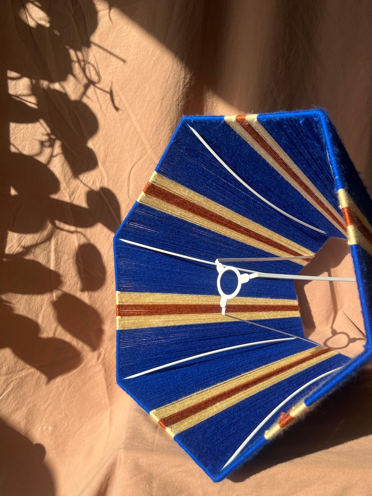 A Slice Shades handmade octagonal fabric lampshade with a royal blue base and diagonal stripes of lemon and rust orange, shown on a beige fabric background with leaf shadows.