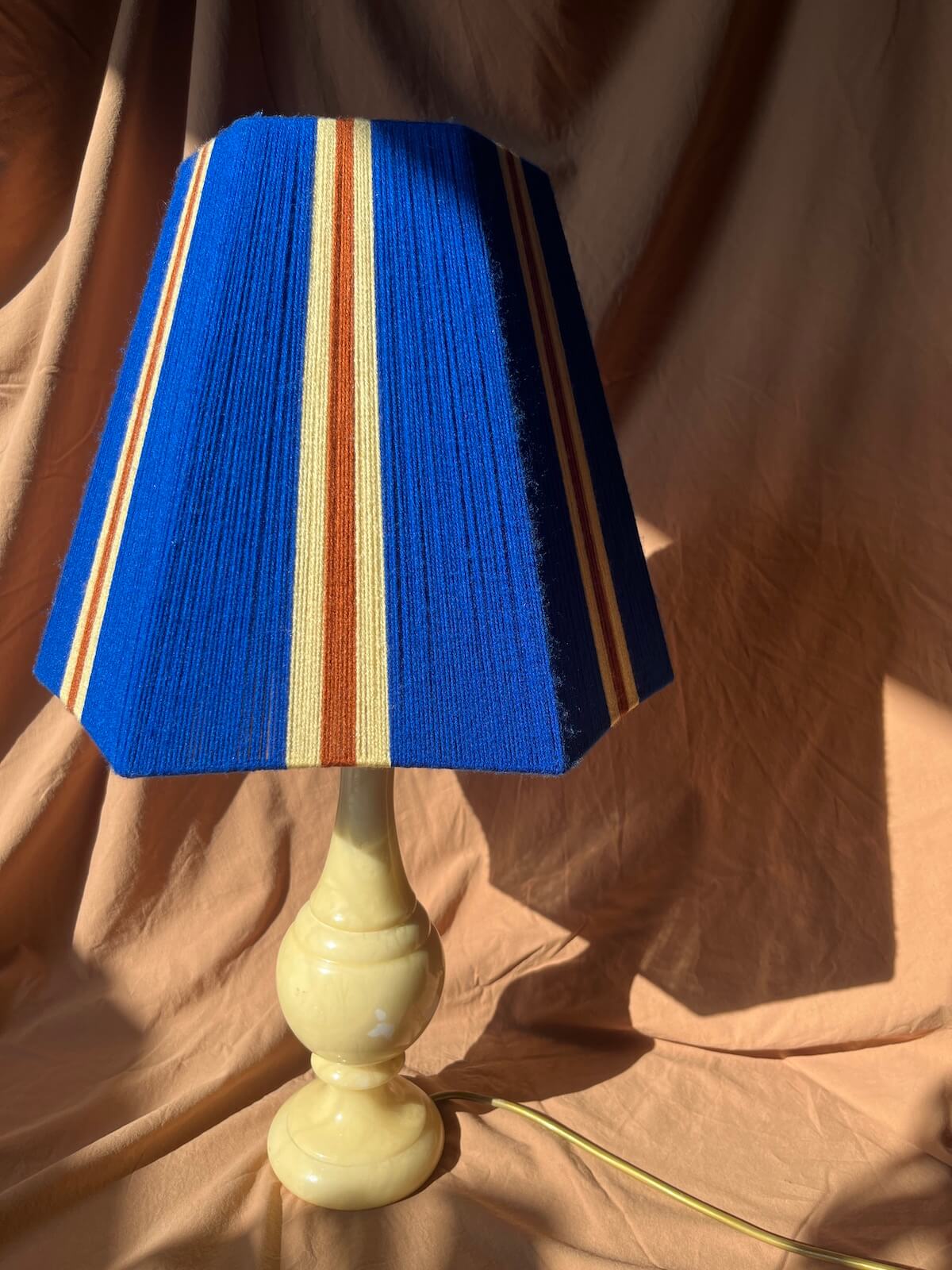 A table lamp with a bSlice Shades yarn lampshade featuring vertical stripes in butter yellow and cobalt blue  mounted on a glossy cream-coloured turned base against a draped tan fabric backdrop.