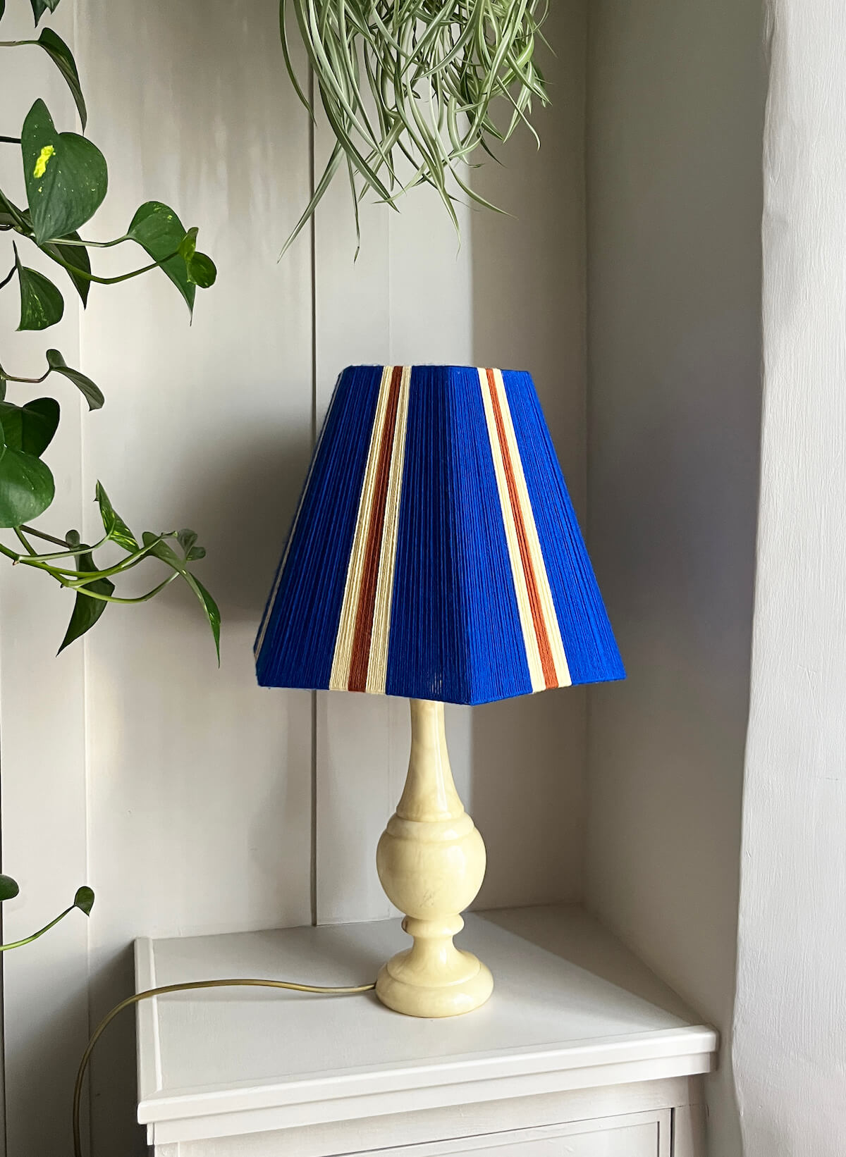 A Slice Shades yarn lampshade in royal blue with vertical yellow and rust-orange stripes, set on a white side table against a panelled wall with houseplants.