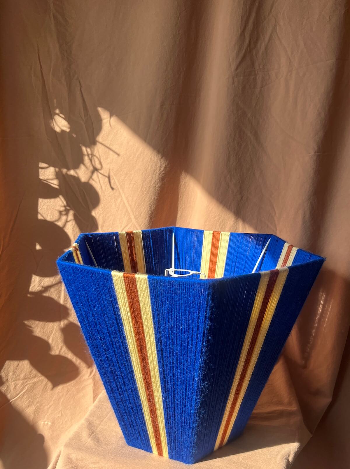 A Slice Shades yarn lampshade with vertical stripes of yellow, rust and royal blue set against a draped beige fabric backdrop with leaf shadows.
