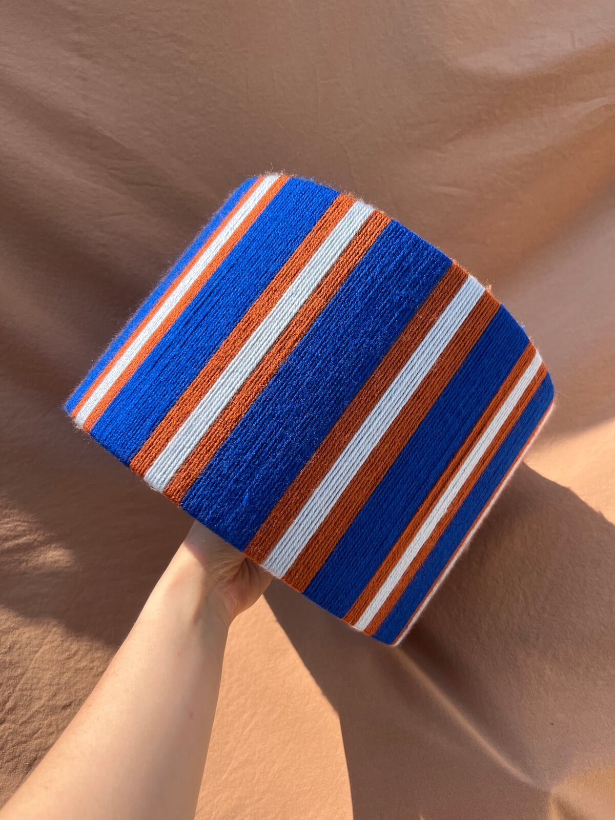 A Slice Shades yarn drum lampshade  with vertical blue, orange, and light blue stripes held by a hand against a draped tan fabric background.