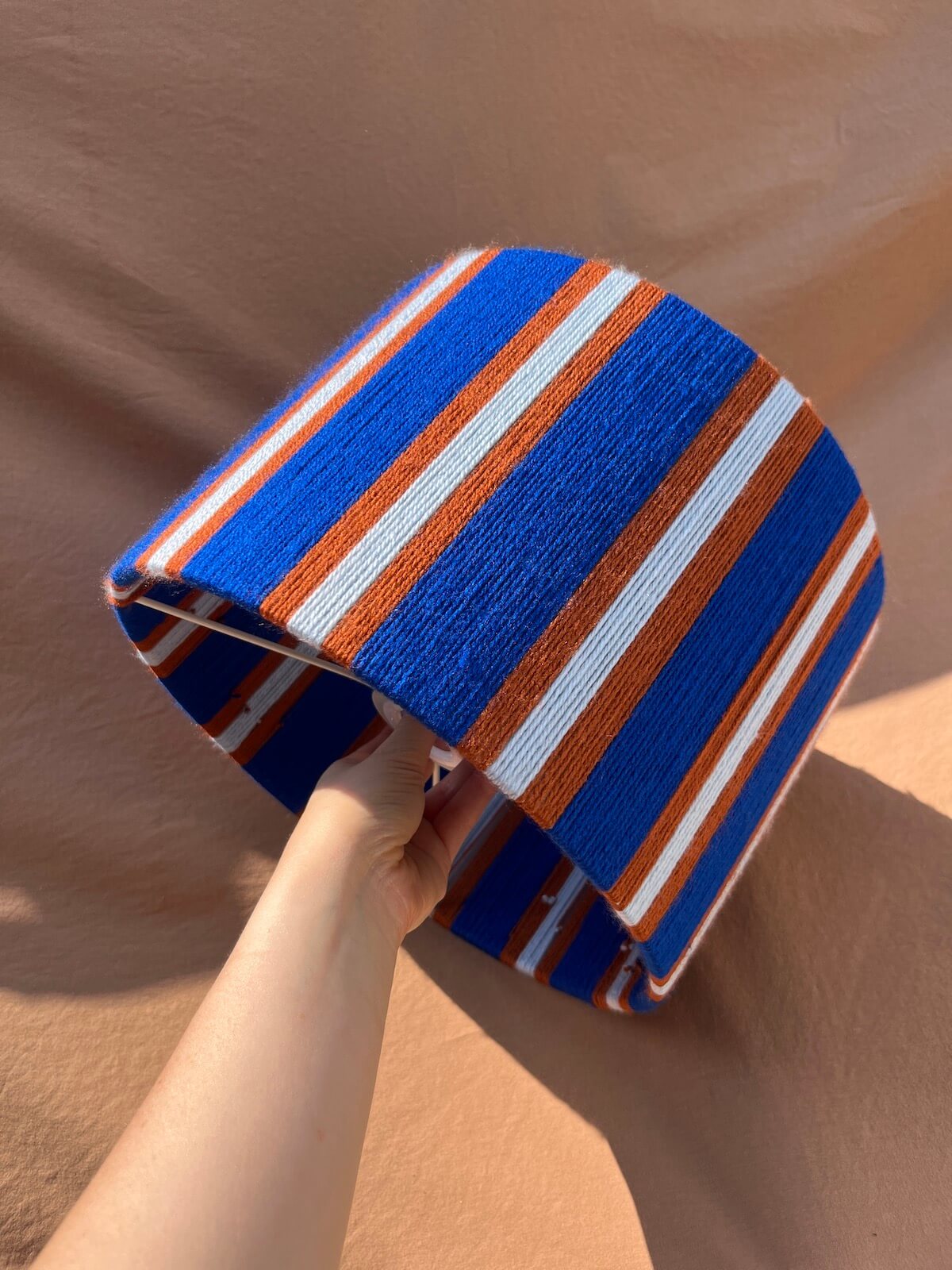 A hand holding a A Slice Shades yarn drum lampshade  with vertical blue, orange, and white stripes set against a warm, textured tan background.