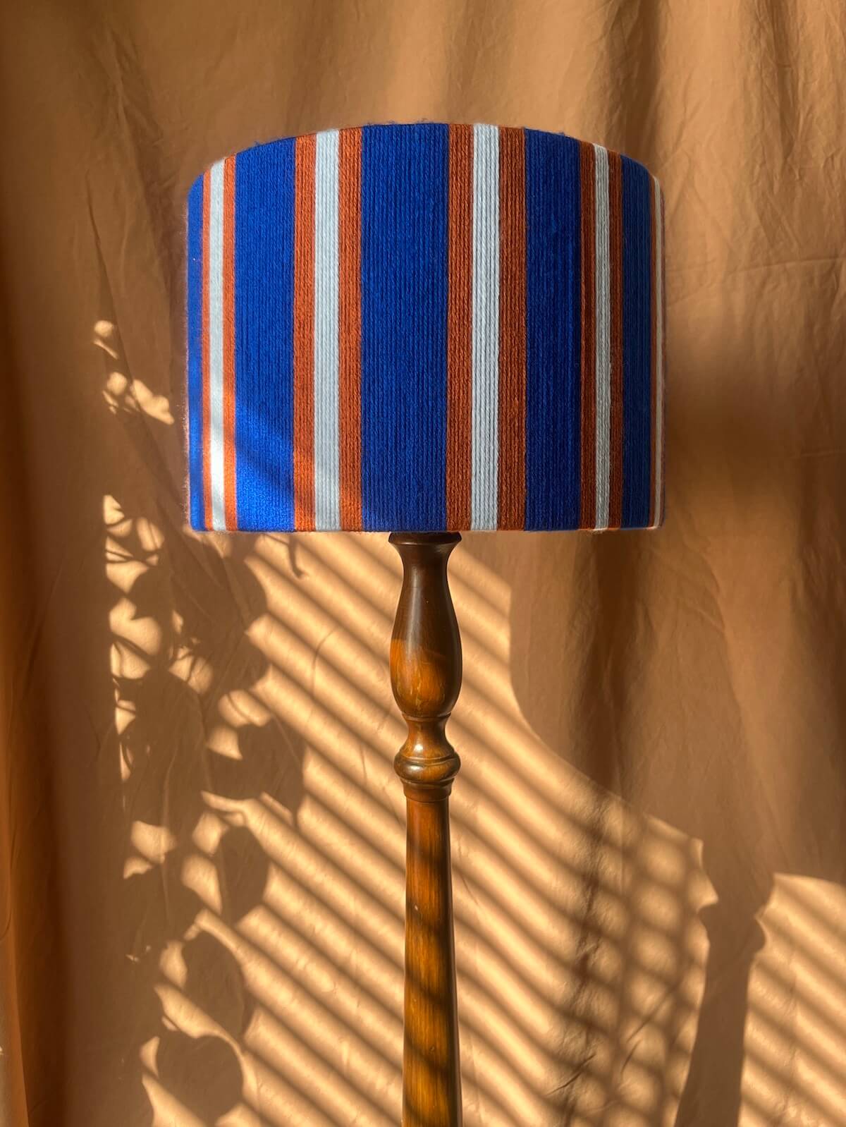 A floor lamp with a Slice Shades yarn drum lampshade  featuring vertical stripes in cobalt blue, rust orange, and off-white, mounted on a turned wooden pole. The lamp is photographed against a draped tan fabric backdrop with dramatic leaf shadows cast across it.