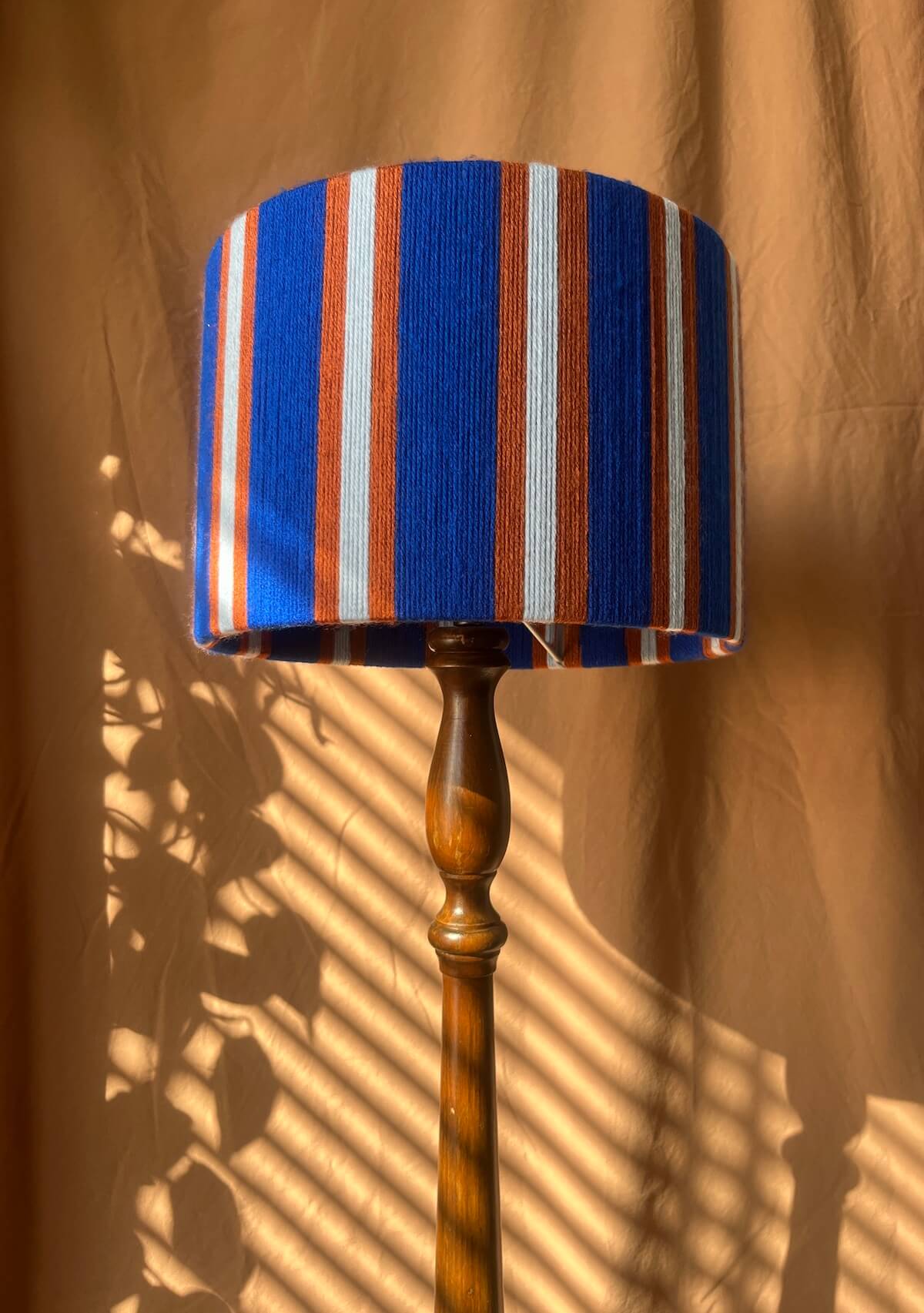 A floor lamp with a Slice Shades yarn drum lampshade featuring vertical stripes in royal blue, rust orange, and off-white, mounted on a turned wooden stand against a draped tan fabric backdrop with leaf shadows.