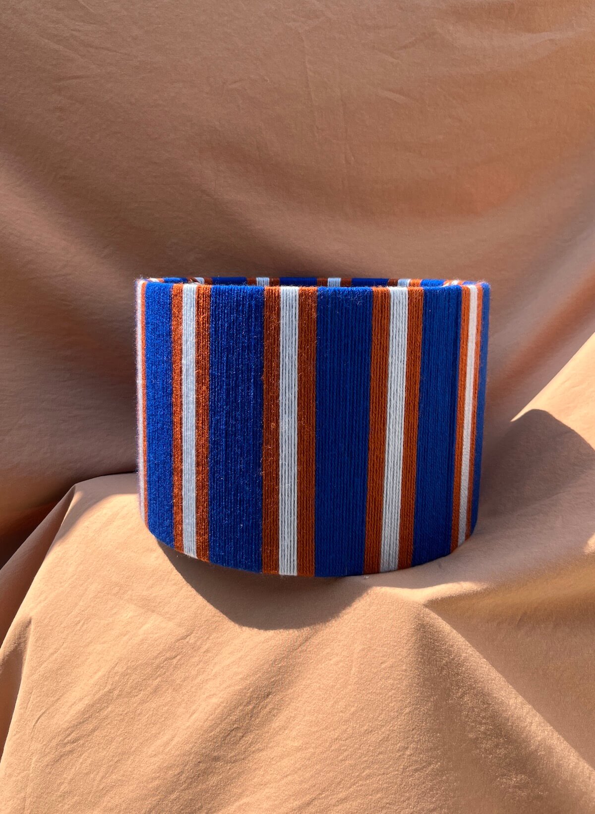 A Slice Shades yarn drum lampshade  with vertical blue, orange, and white stripes with vertical stripes of royal blue, rust orange, and white, displayed on a draped tan fabric background.