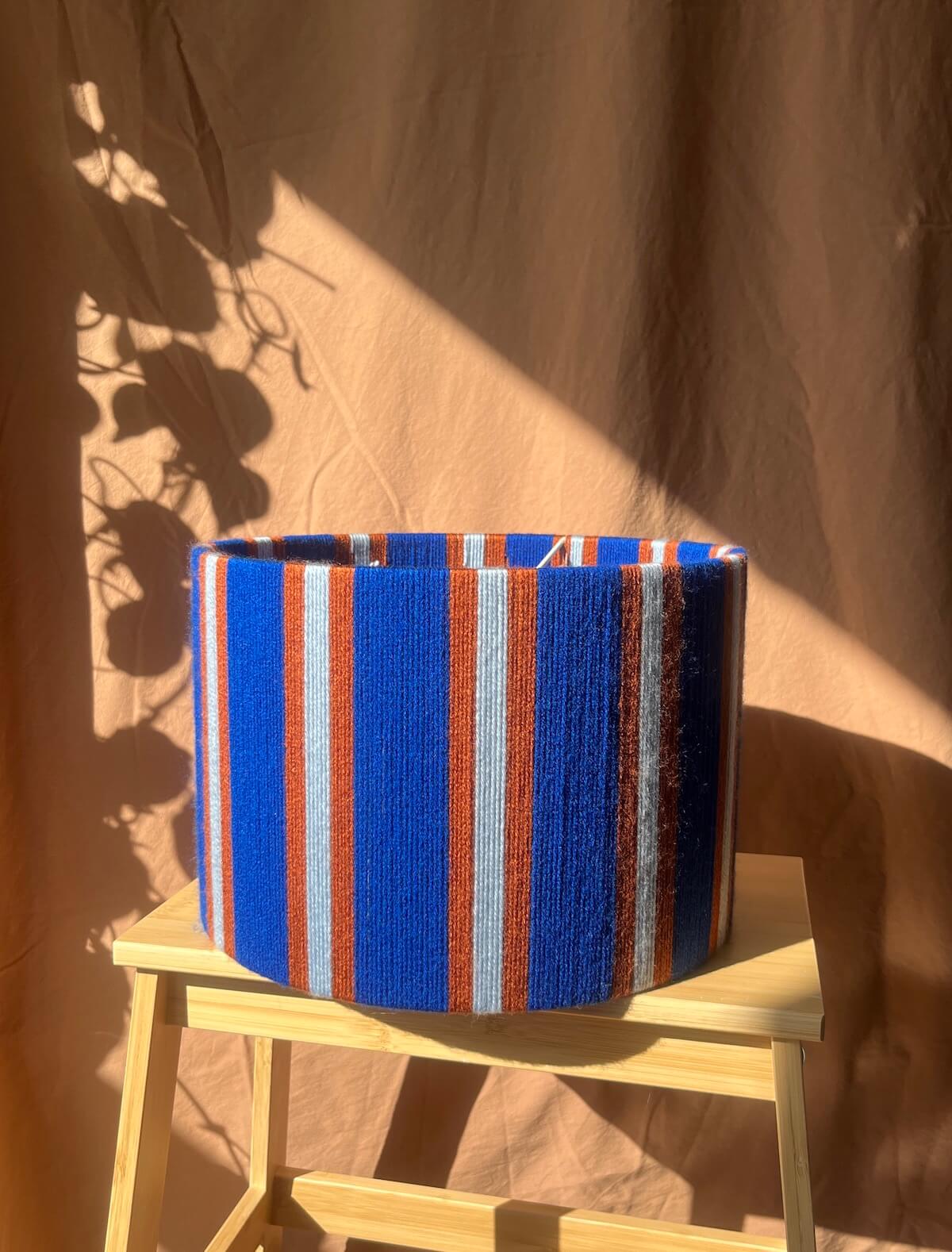 A Slice Shades yarn drum lampshade  with vertical blue, orange, and white stripes, displayed on a small wooden stool against a draped brown fabric backdrop with leaf shadows.