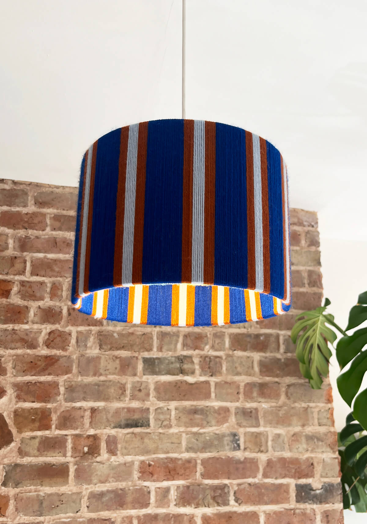 A drum-shaped A Slice Shades yarn drum lampshade with vertical blue, orange, and white stripes hanging against a brick wall background.